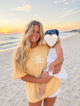 Load image into Gallery viewer, A woman and a child on a sandy beach at sunset. The woman wears a pale yellow oversized graphic T-shirt with the text 'KINDA CHIC TO RAISE TINY HUMANS' in white.
