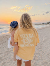 Load image into Gallery viewer, A woman and a child standing on a sandy beach at sunset, viewed from behind. The woman is wearing a pale yellow graphic T-shirt with the text 'KINDA CHIC TO RAISE TINY HUMANS' in white, and the child is wearing a blue bandana.
